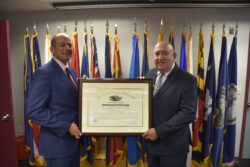 Acting Director Craig Brown standing on the left, Greg Adinolfi standing to the right holding framed certificate in front of flags.